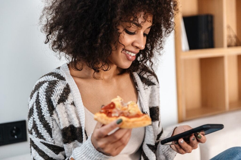 Pretty young african woman indoors at home eat pizza.
